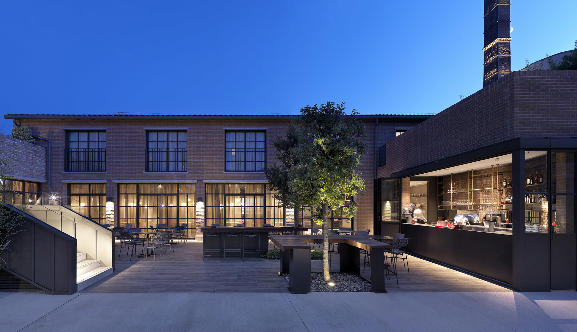 Industrial brick building courtyard at dusk with warm outdoor lighting illuminating trees, dining area, and open bar terrace.