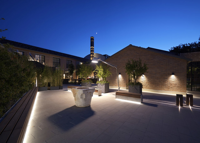 Industrial brick courtyard at dusk with warm LED ground lighting, wall-mounted fixtures, bollards, and a large articulated floor lamp among trees.