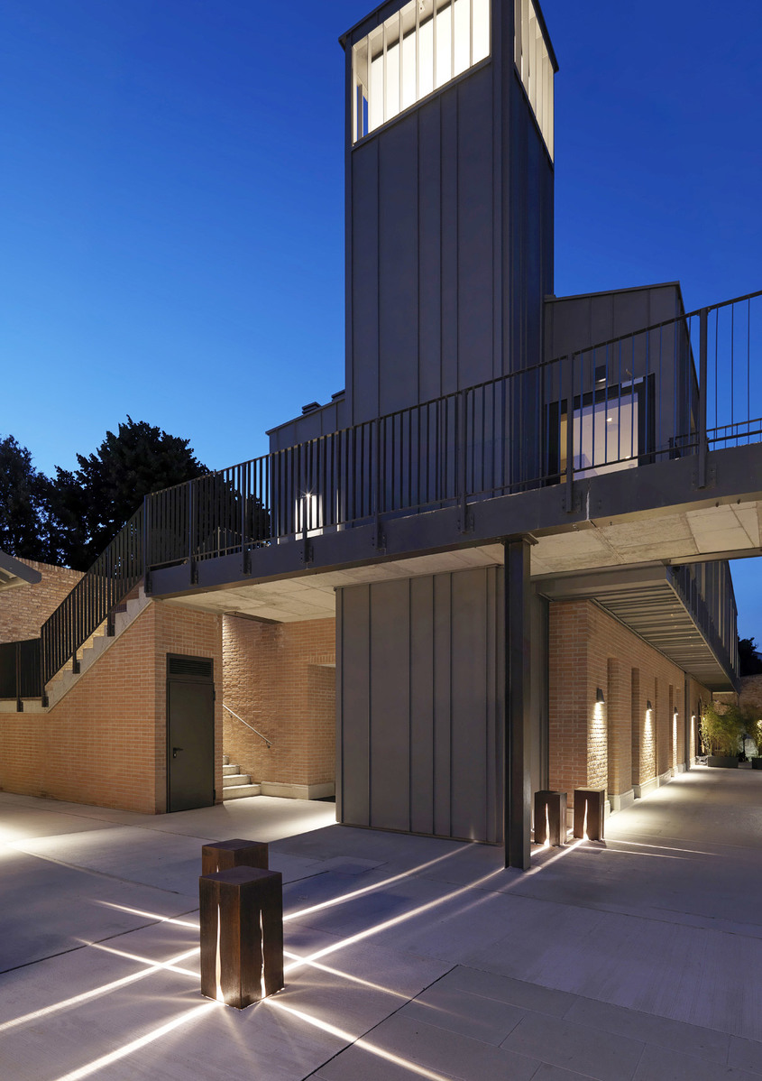 Outdoor courtyard at dusk with Artemide bollard lights casting dramatic cross-shaped beams on paved ground near a brick building