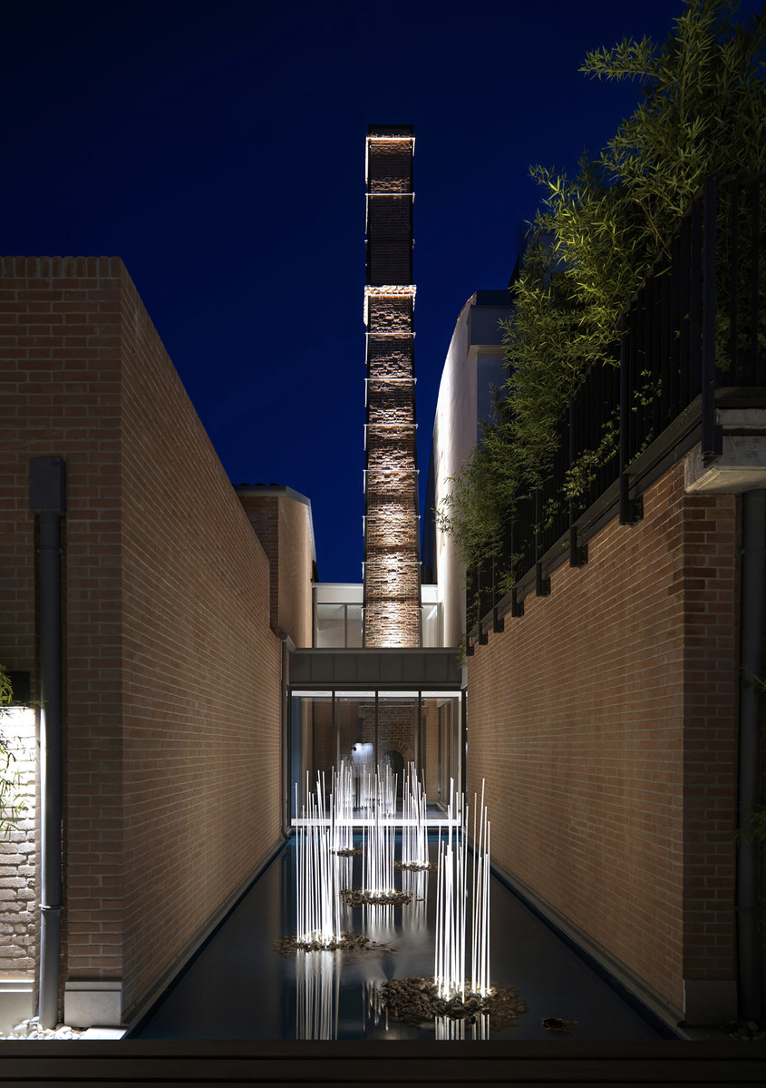 Nighttime courtyard with illuminated brick chimney tower, reflective pool, and vertical light installations between brick walls