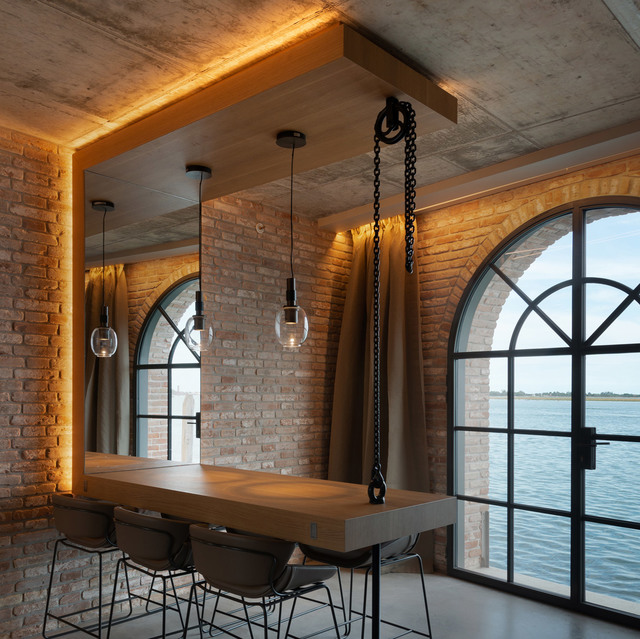 Industrial dining area with pendant glass globe lights over a wooden bar table, warm LED cove lighting, brick walls, and arched windows overlooking water.