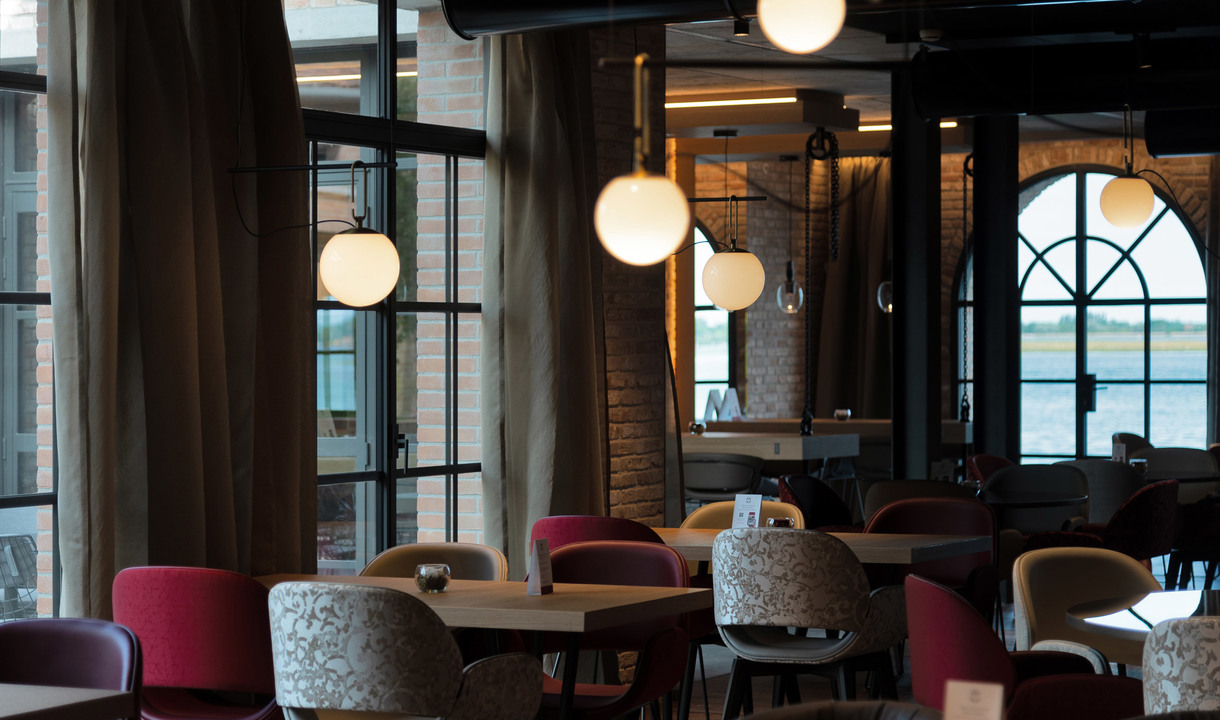 Restaurant interior with spherical white glass pendant lamps hanging over wooden tables and colorful chairs, large windows with waterfront view