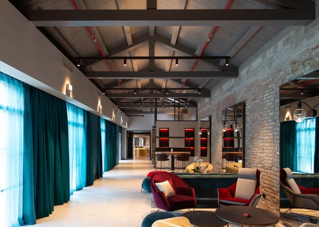 Modern hotel lounge with vaulted ceiling, track lighting, teal curtains, exposed brick walls, and colorful seating areas.