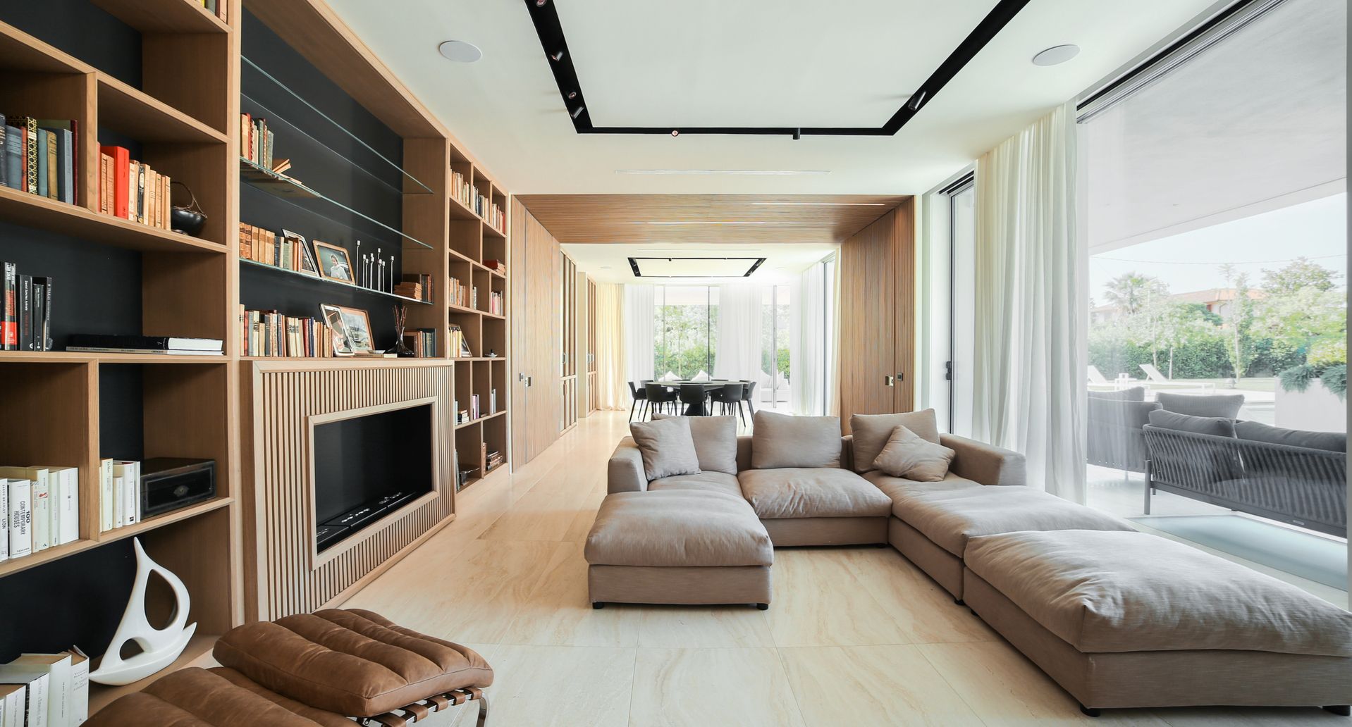 Modern living room with large sectional sofa, built-in wooden bookcase, recessed ceiling lights, and open view to dining area and terrace