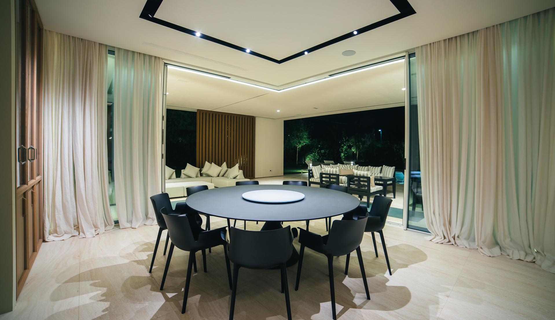 Modern dining room with round dark table, black chairs, and geometric ceiling recess with integrated LED lighting opening to outdoor terrace at night