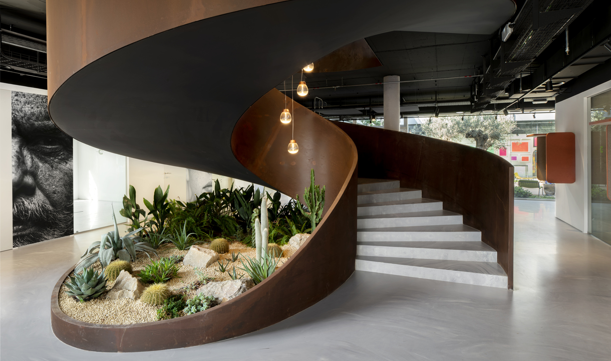 Spiral staircase with Corten steel base enclosing a desert garden, illuminated by three amber pendant bulbs hanging from above.
