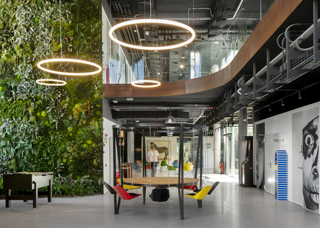 Modern office atrium with Artemide Halo ring pendant lights over a collaborative seating area beside a lush green living wall.
