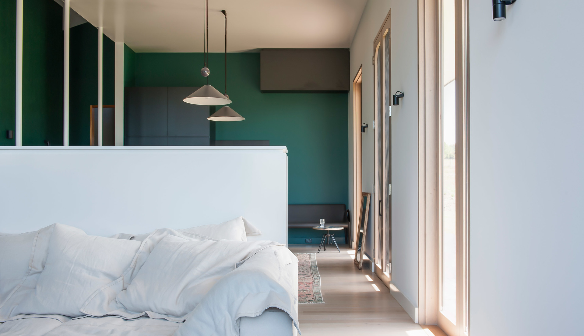 Modern bedroom with green accent wall featuring two conical pendant lamps hanging above a white bed and small seating area