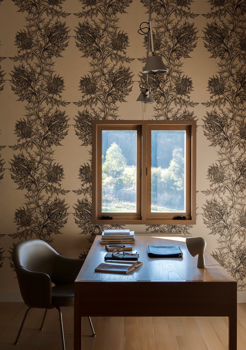 Wall-mounted adjustable metal task lamp above a wooden desk with books, in a study with floral wallpaper and a window