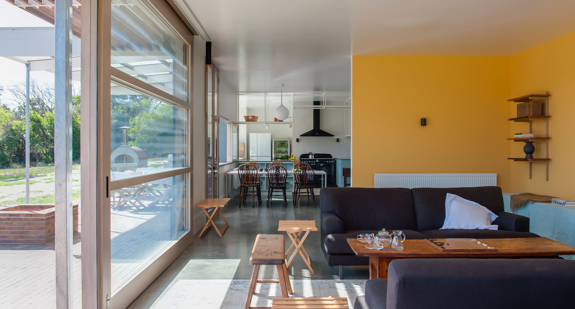 Open-plan living and dining area with large glass sliding doors, dark sofa, wooden coffee table, and a white pendant lamp over the kitchen.