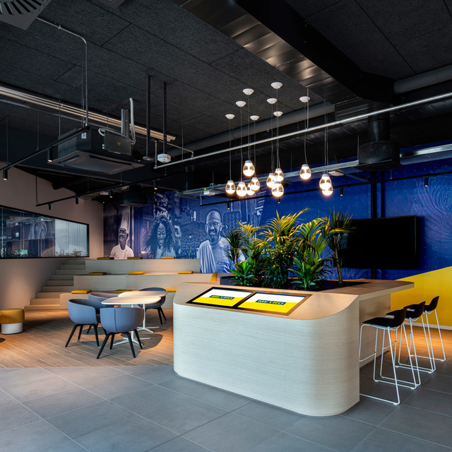 Modern office lounge with Artemide pendant lights hanging over a curved reception desk, plants, blue mural, and yellow accents.