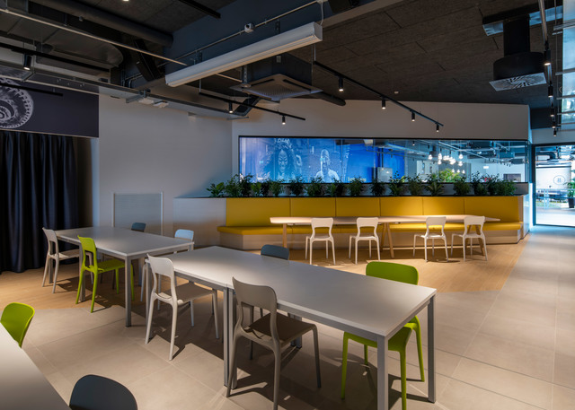 Modern office cafeteria with track lighting on dark ceiling, white tables, colorful chairs, yellow booth seating, and green plants.