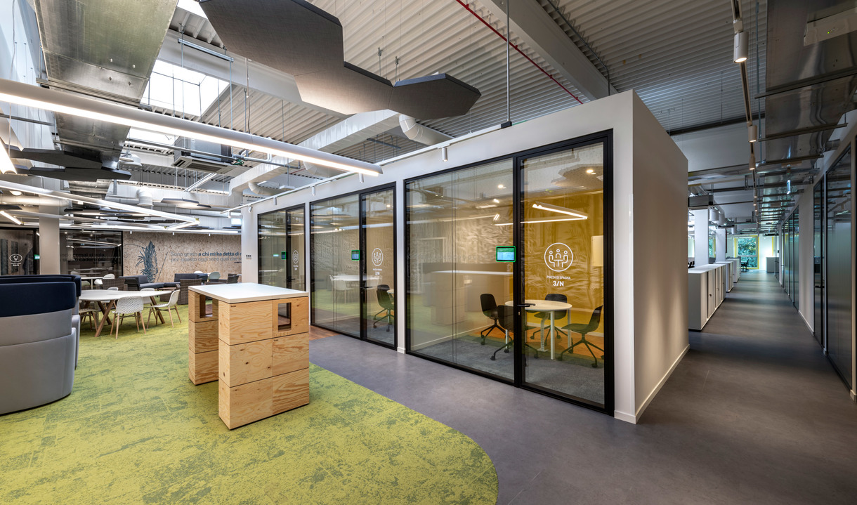 Modern open-plan office with glass meeting pod, green carpet, wooden storage units, and industrial ceiling with pendant lighting.