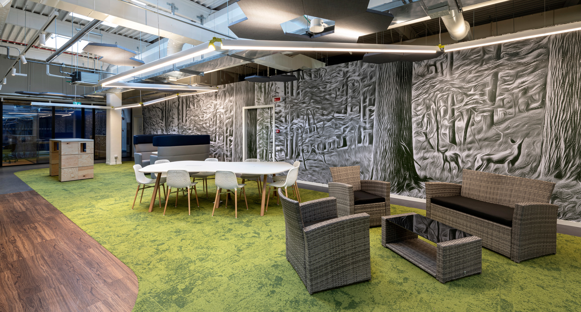 Modern office lounge with Artemide linear ceiling lights, wicker seating, green moss carpet, and a textured forest-themed wall mural.