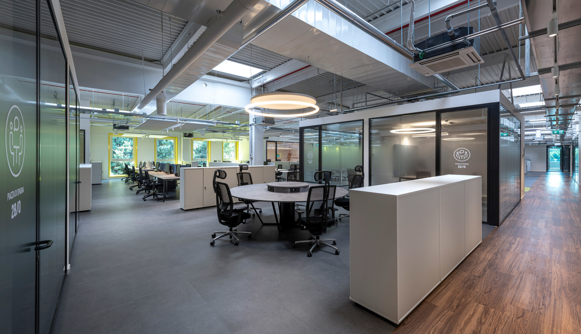 Modern open-plan office with Artemide circular pendant light, glass-partitioned meeting rooms, workstations, and industrial-style exposed ceiling.