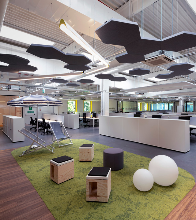 Modern open-plan office with hexagonal acoustic ceiling panels, white pendant lights, green carpet area with wooden stools and spherical floor lamps
