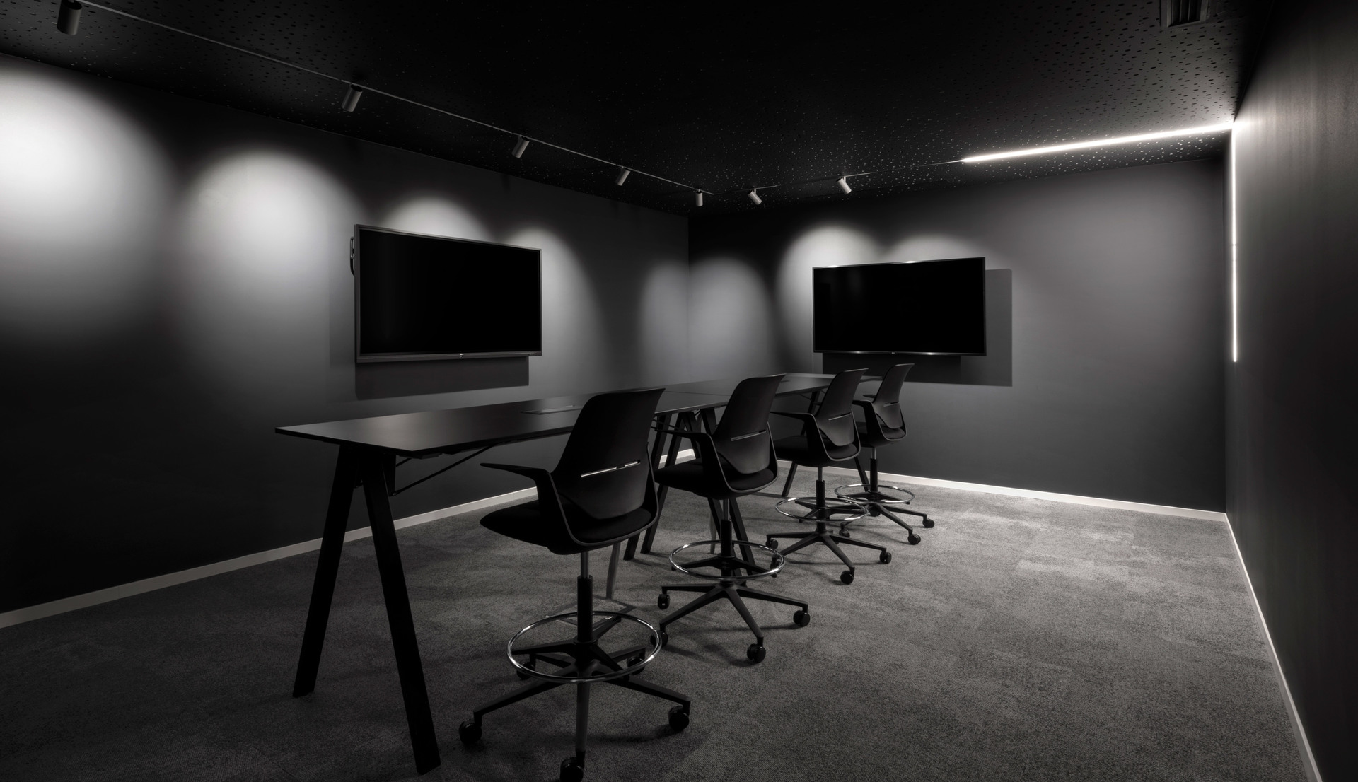 Dark meeting room with track spotlights, a linear LED strip on the wall, long black desk, ergonomic chairs, and two wall-mounted screens.