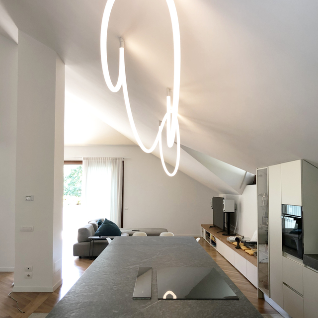 Artemide Alphabet of Light large pendant forming a cursive letter shape, illuminating a modern open-plan kitchen with sloped ceiling