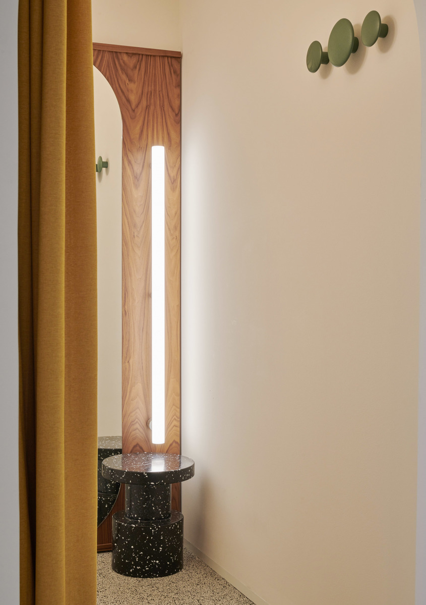 Slim vertical floor lamp with glowing white tube beside dark terrazzo side table, warm mustard curtain, and olive green wall hooks.