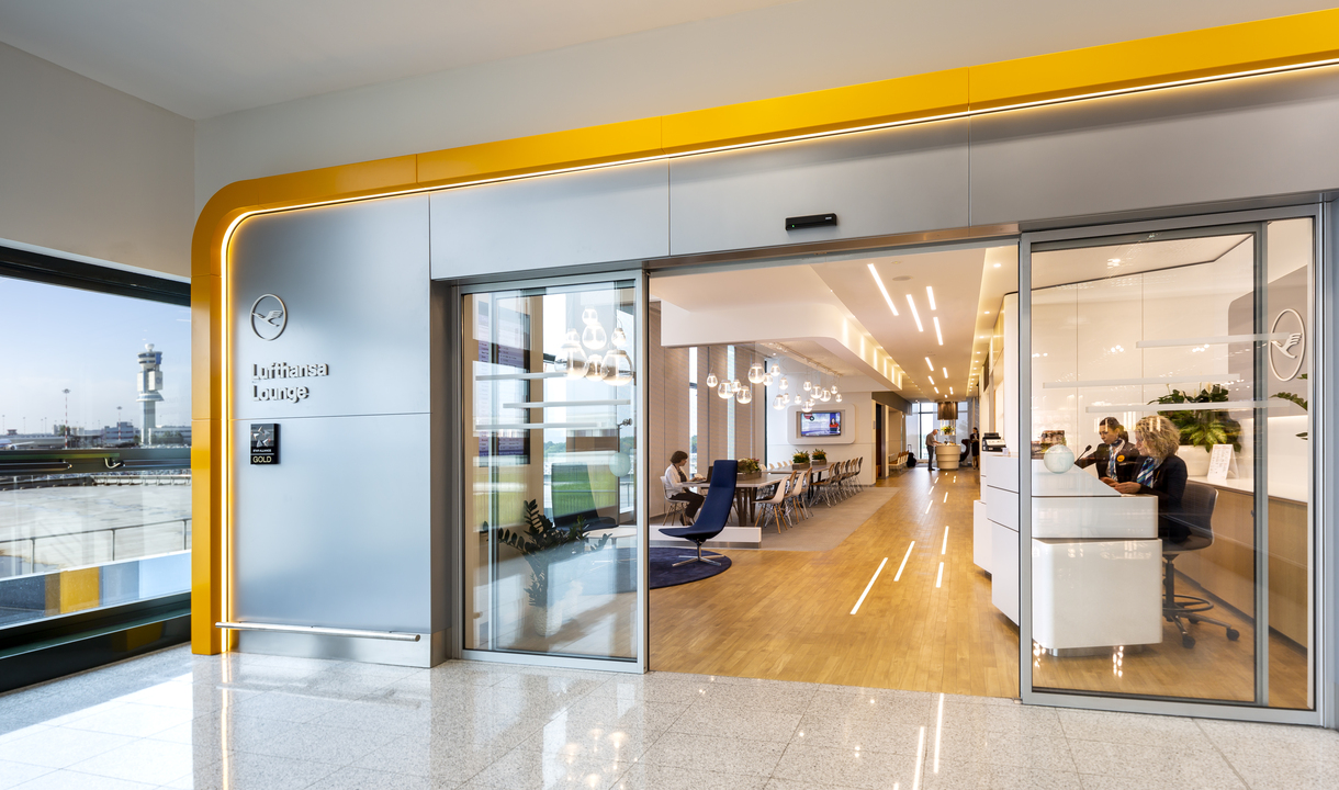 Modern airport lounge entrance with glass doors, yellow accents, and Artemide recessed ceiling lights illuminating a seating area.