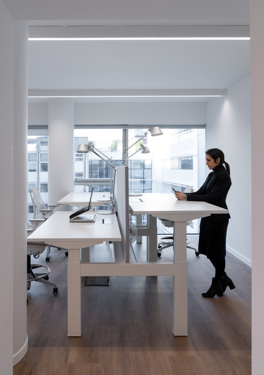 Modern office workspace with Artemide Tolomeo desk lamps on white standing desks, linear ceiling light, and a woman working standing up.