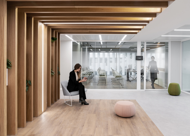 Modern office lounge with warm wood-paneled corridor, linear ceiling lights, glass partitions, and a woman seated using a tablet.