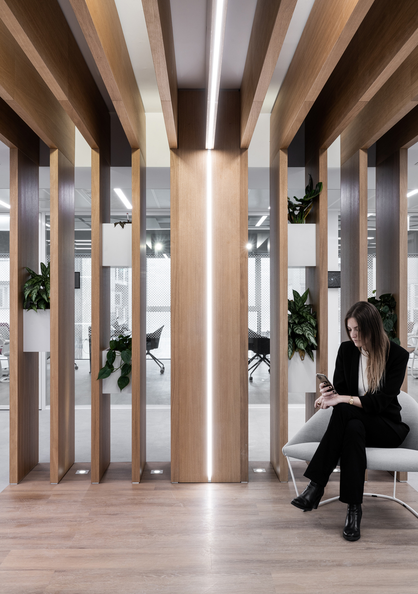 Modern office lounge with wooden columns featuring integrated linear LED lighting, a woman seated checking her phone, and green plants.