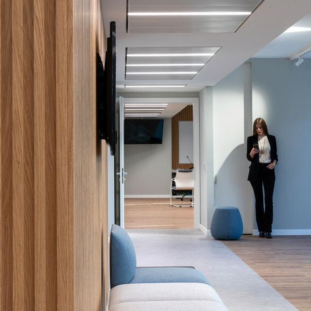 Modern office corridor with recessed linear LED ceiling lights, wood-paneled wall, seating area, and a woman checking her phone.