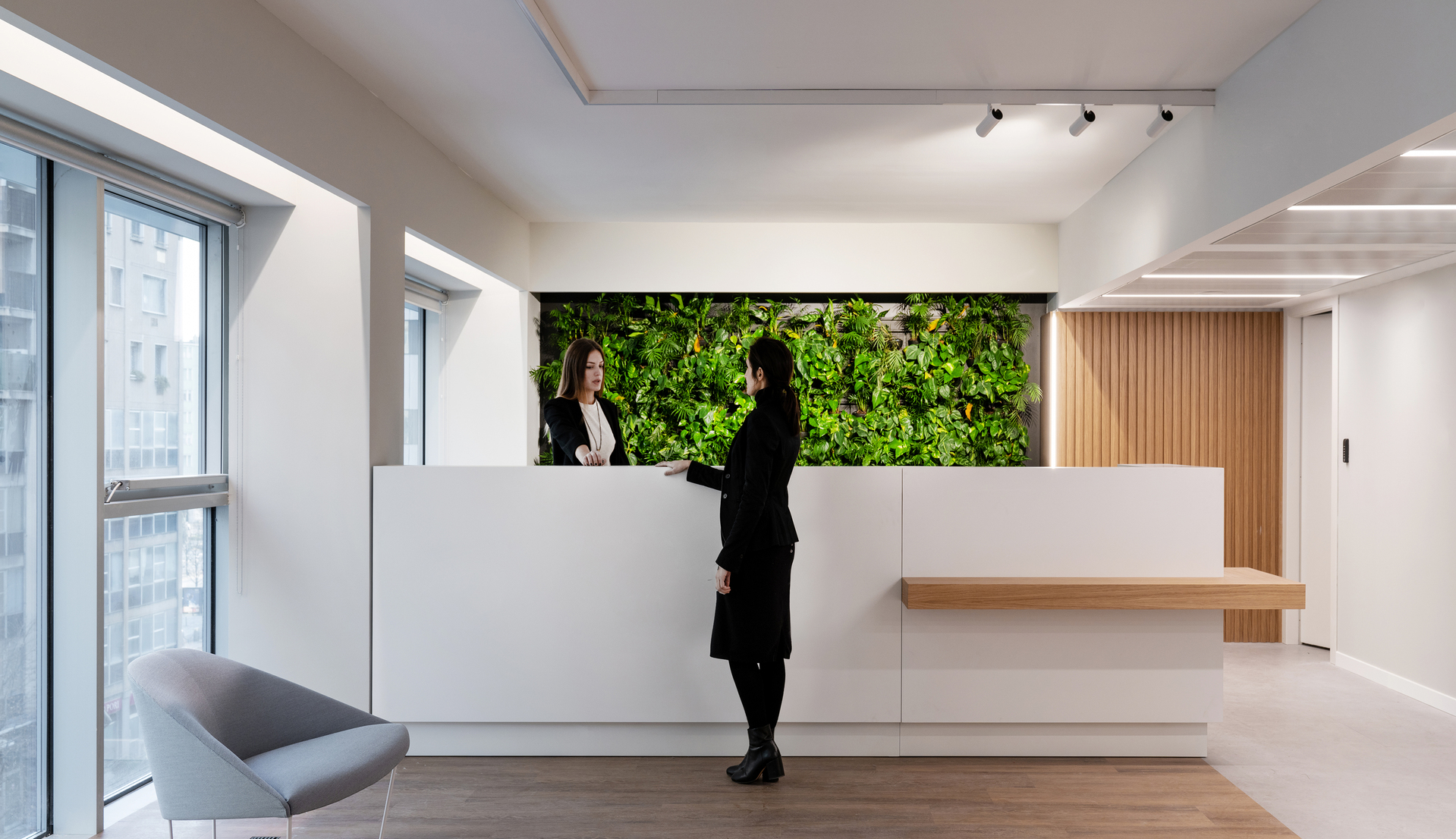 Modern office reception with Artemide track and linear lights, two women at white desk, living green wall backdrop.