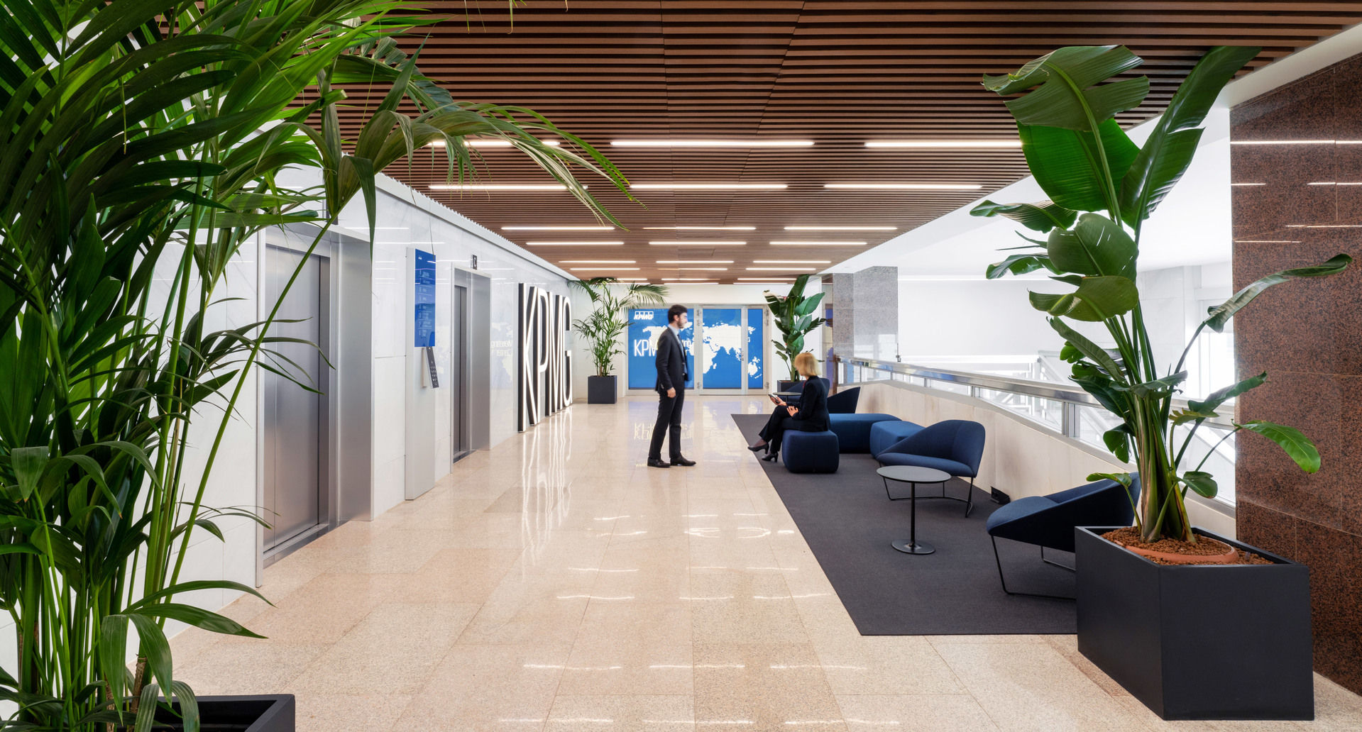 Modern office corridor with linear ceiling lights, tropical plants, lounge seating area, and two people conversing near elevator doors.