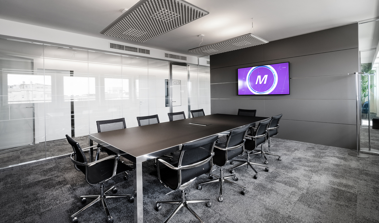 Modern conference room with a long dark table, mesh chairs, recessed ceiling lighting, and a wall-mounted display screen.