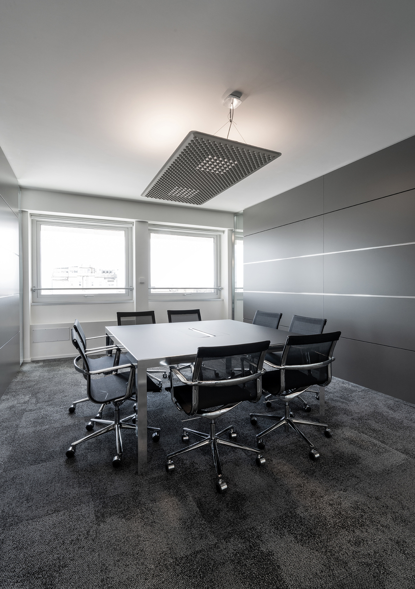 Modern conference room with a large square pendant light fixture illuminating a rectangular table surrounded by six black office chairs