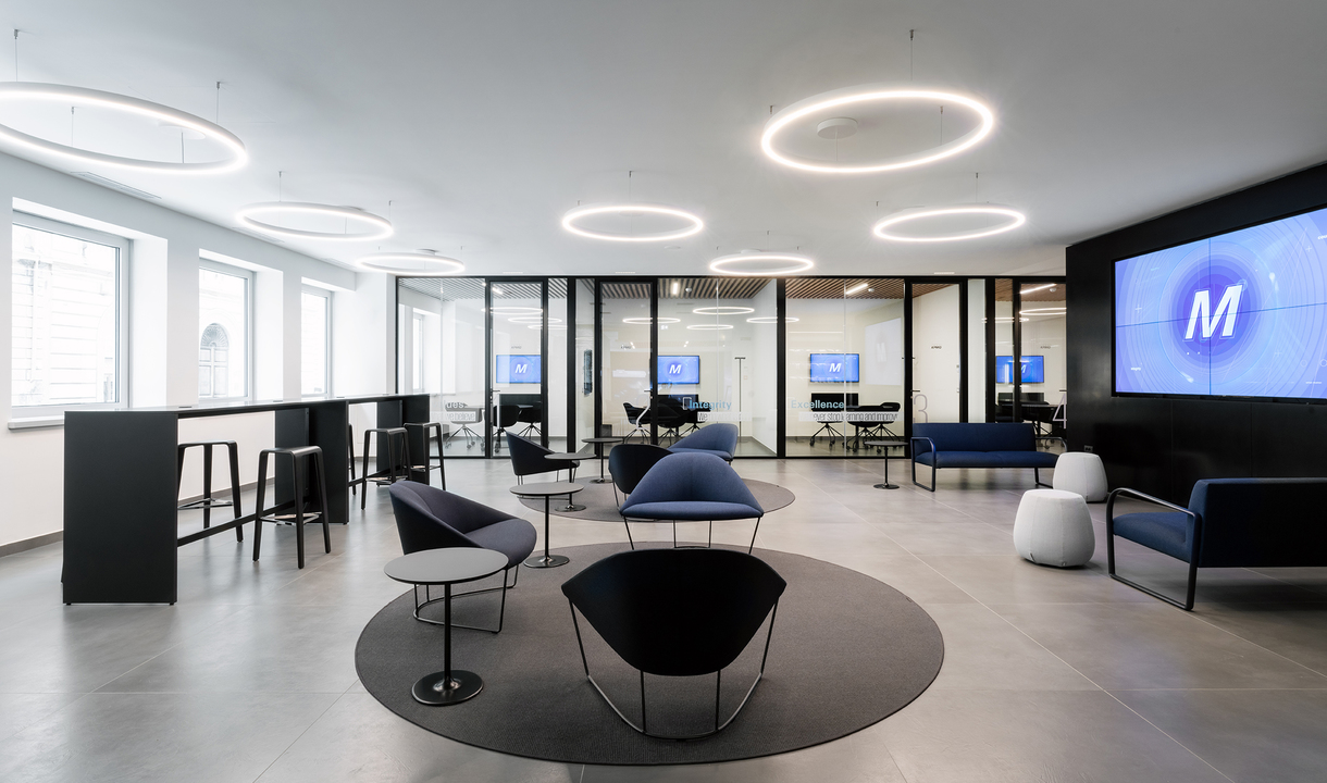 Modern office lounge with circular LED ring pendant lights illuminating a collaborative seating area with dark chairs and glass-partitioned meeting rooms.
