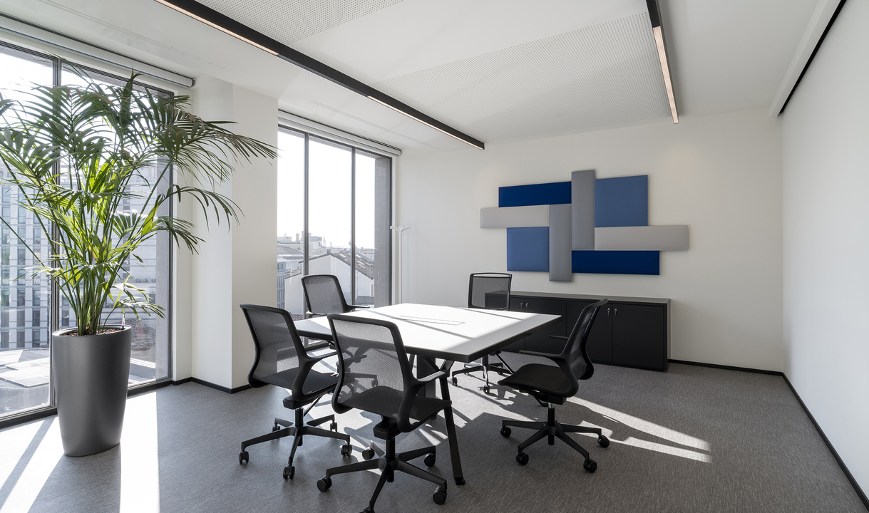 Modern meeting room with recessed linear ceiling lights, white table, black mesh chairs, acoustic panels, and a large indoor plant.