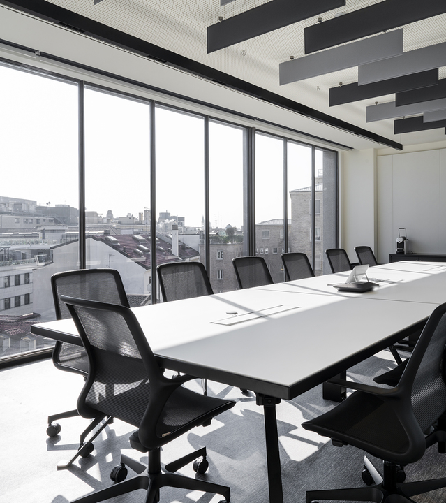 Modern conference room with long white table, black mesh chairs, floor-to-ceiling windows, and dark acoustic panels on ceiling.