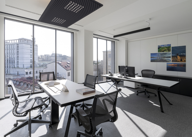 Modern office with white desks, mesh chairs, large windows, and a perforated black ceiling panel providing ambient lighting.