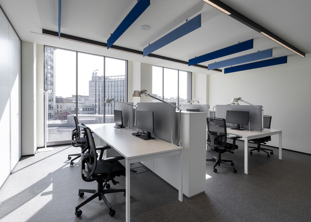Modern open-plan office with white desks, task chairs, desktop monitors, desk lamps, large windows, and blue acoustic panels on ceiling.