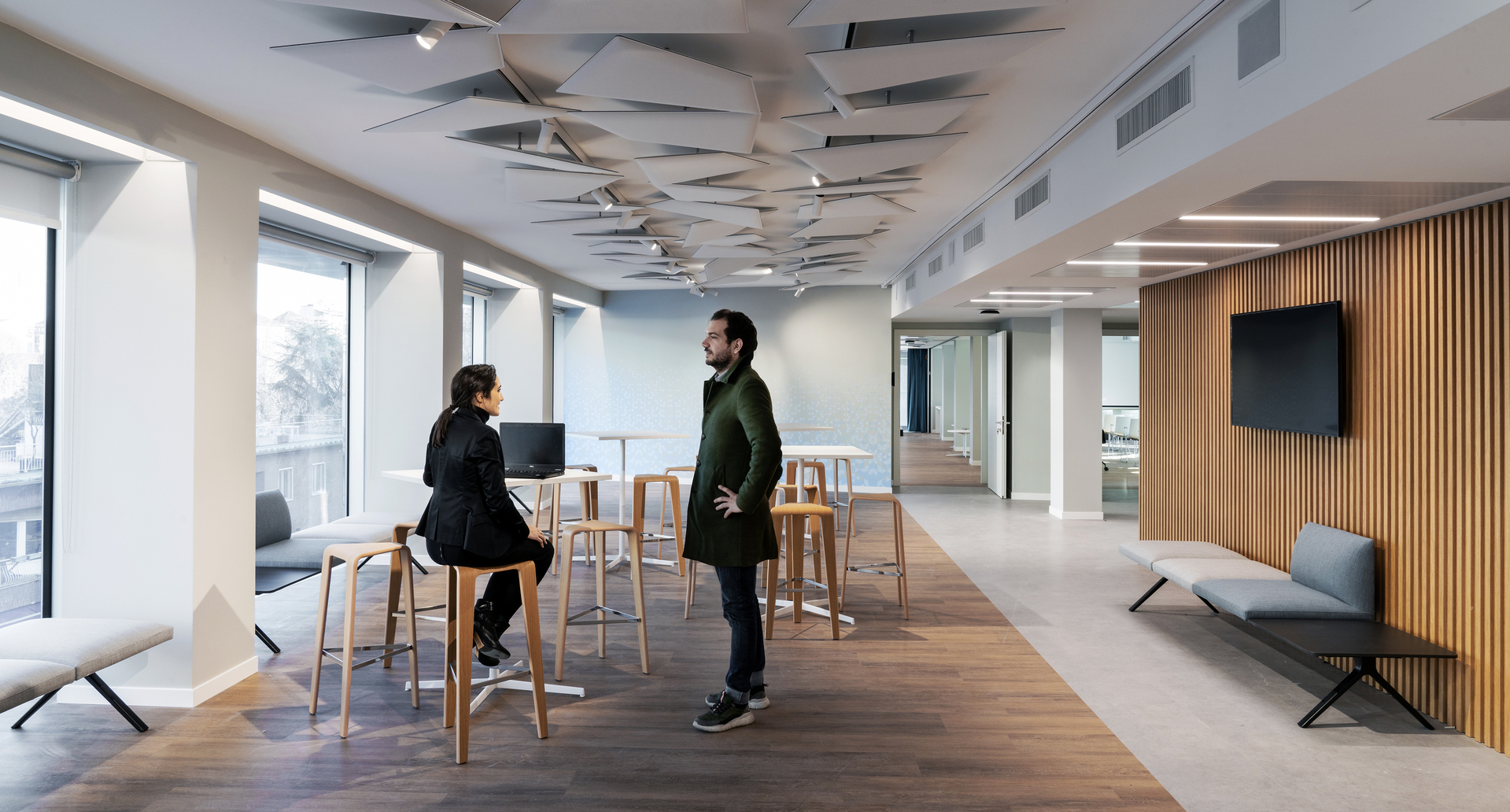 Modern office lounge with two people conversing near high tables, featuring decorative ceiling panels and linear LED lighting above.