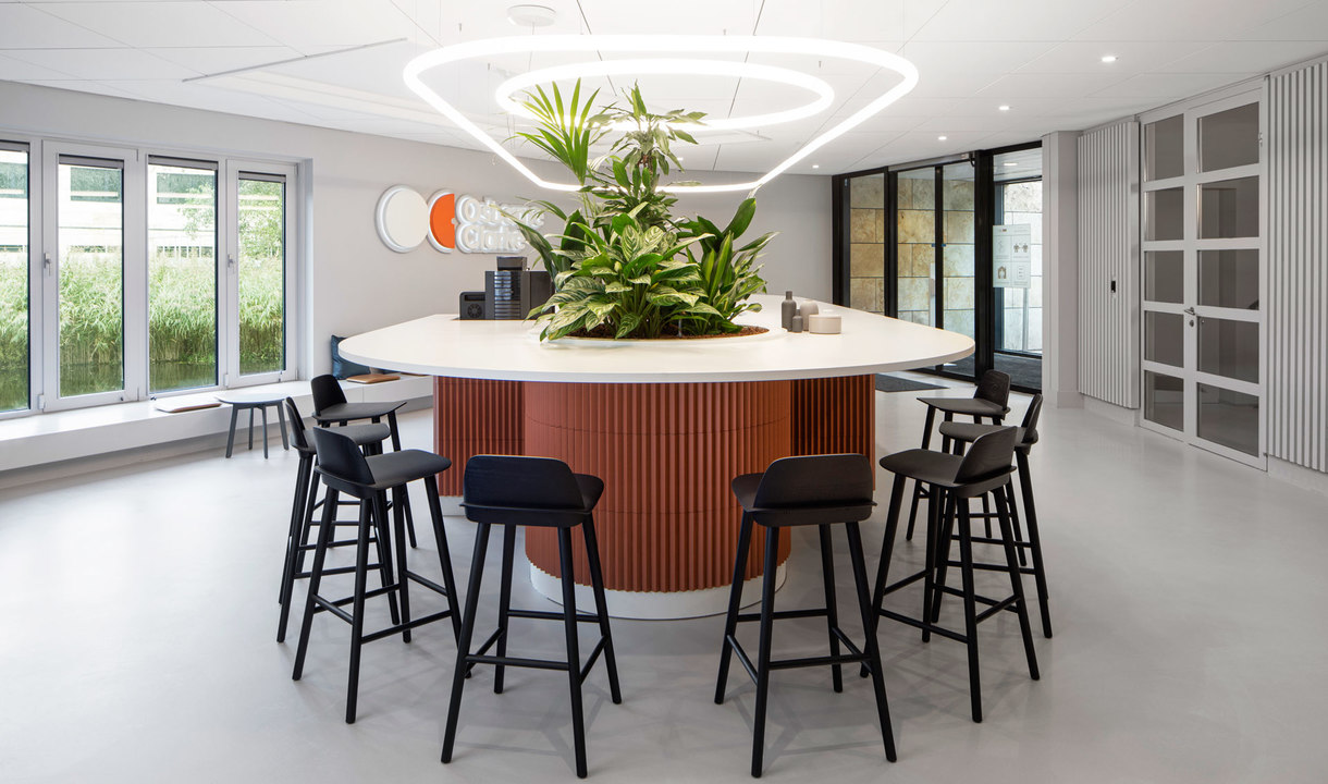 Modern office lounge with triangular Artemide pendant light above a round bar table with plants and black stools.