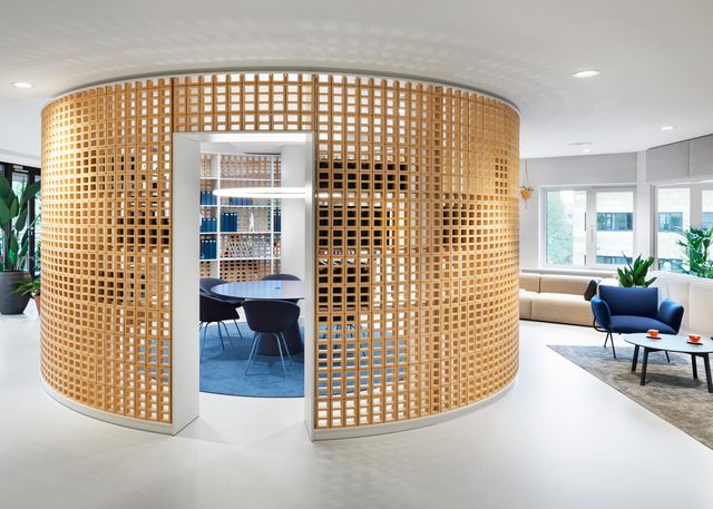 Modern office lounge with curved wooden lattice partition enclosing a meeting room, blue seating, and recessed ceiling lights.