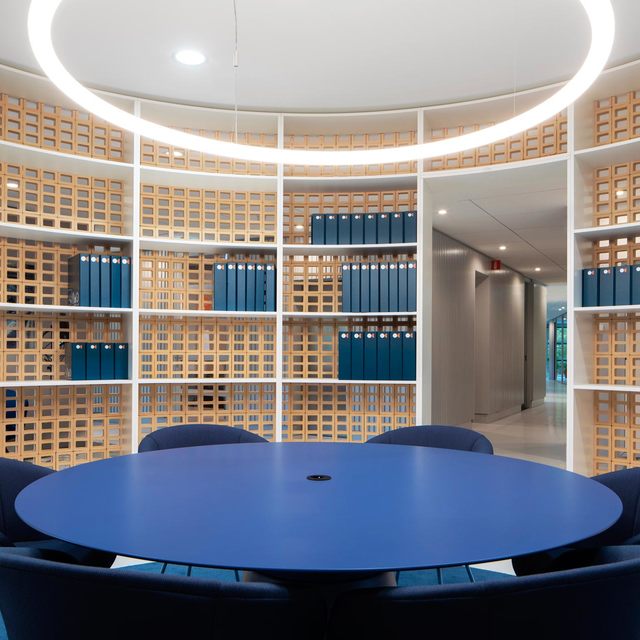 Circular meeting room with round blue table, navy chairs, wood lattice shelving, and an Artemide ring pendant light overhead.