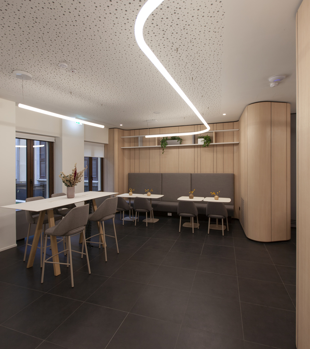 Modern café interior with a curved LED ceiling light strip, bar tables, gray chairs, and a upholstered bench seating area.