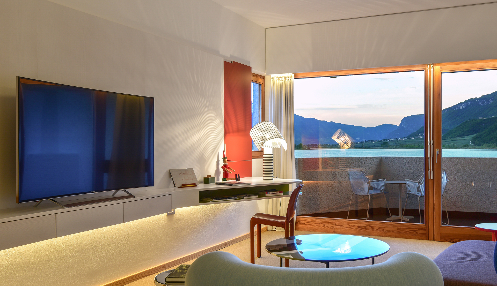 Modern living room with Artemide Tolomeo table lamp on desk, warm LED strip lighting, large window overlooking mountain landscape at dusk