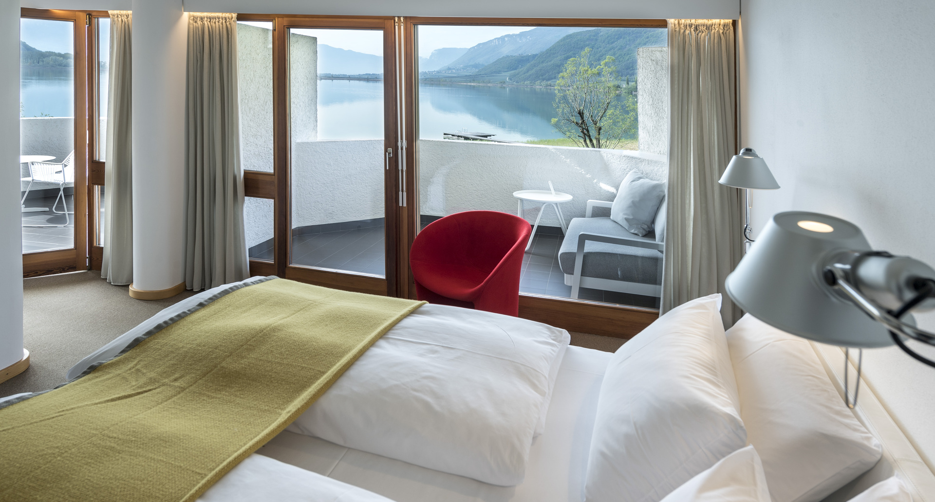 Hotel bedroom with Artemide Tolomeo bedside lamp, white bedding, yellow throw, and balcony overlooking a scenic lake and mountains