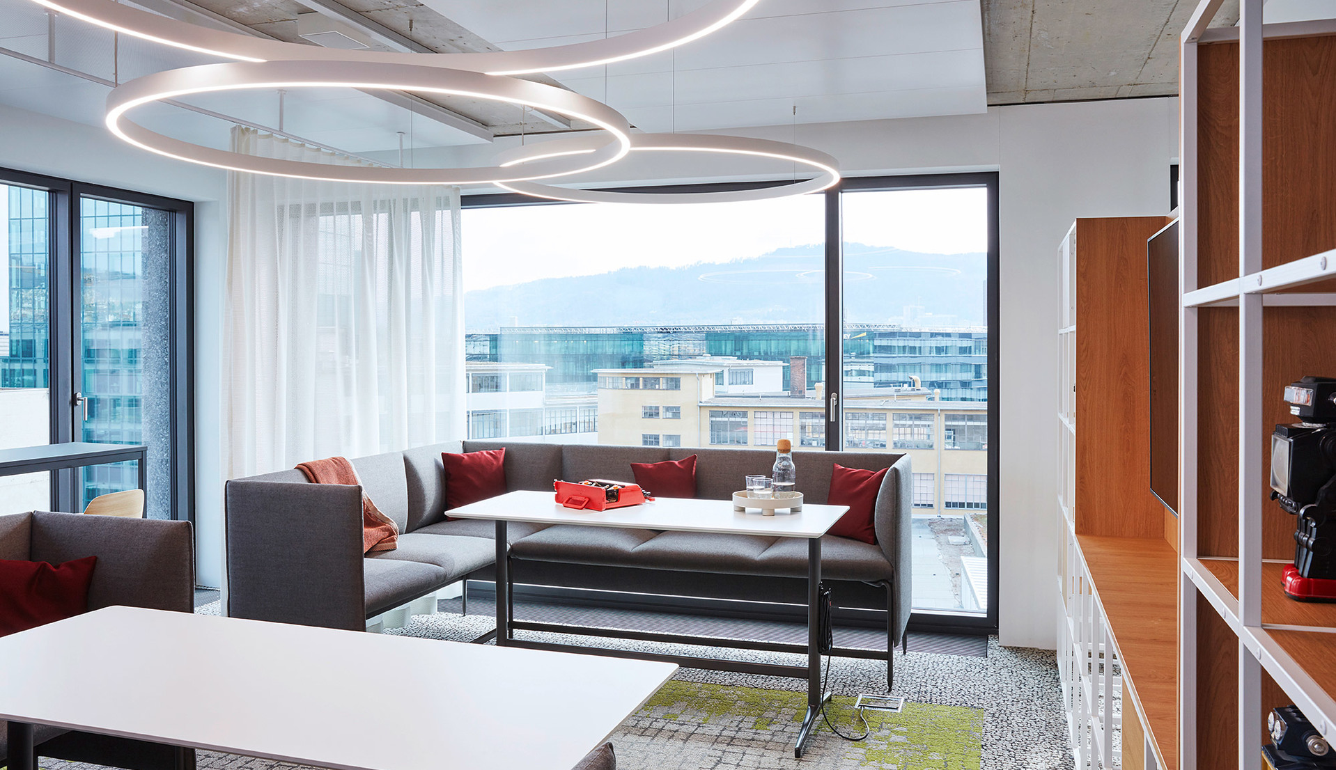 Artemide circular ring pendant light illuminates a modern office lounge with gray sofas, red cushions, and panoramic city views.