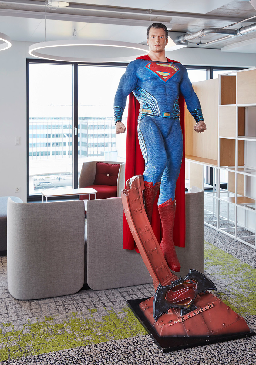 Life-size Superman statue in a modern office lounge with circular ceiling light, grey sofas, and open shelving unit.