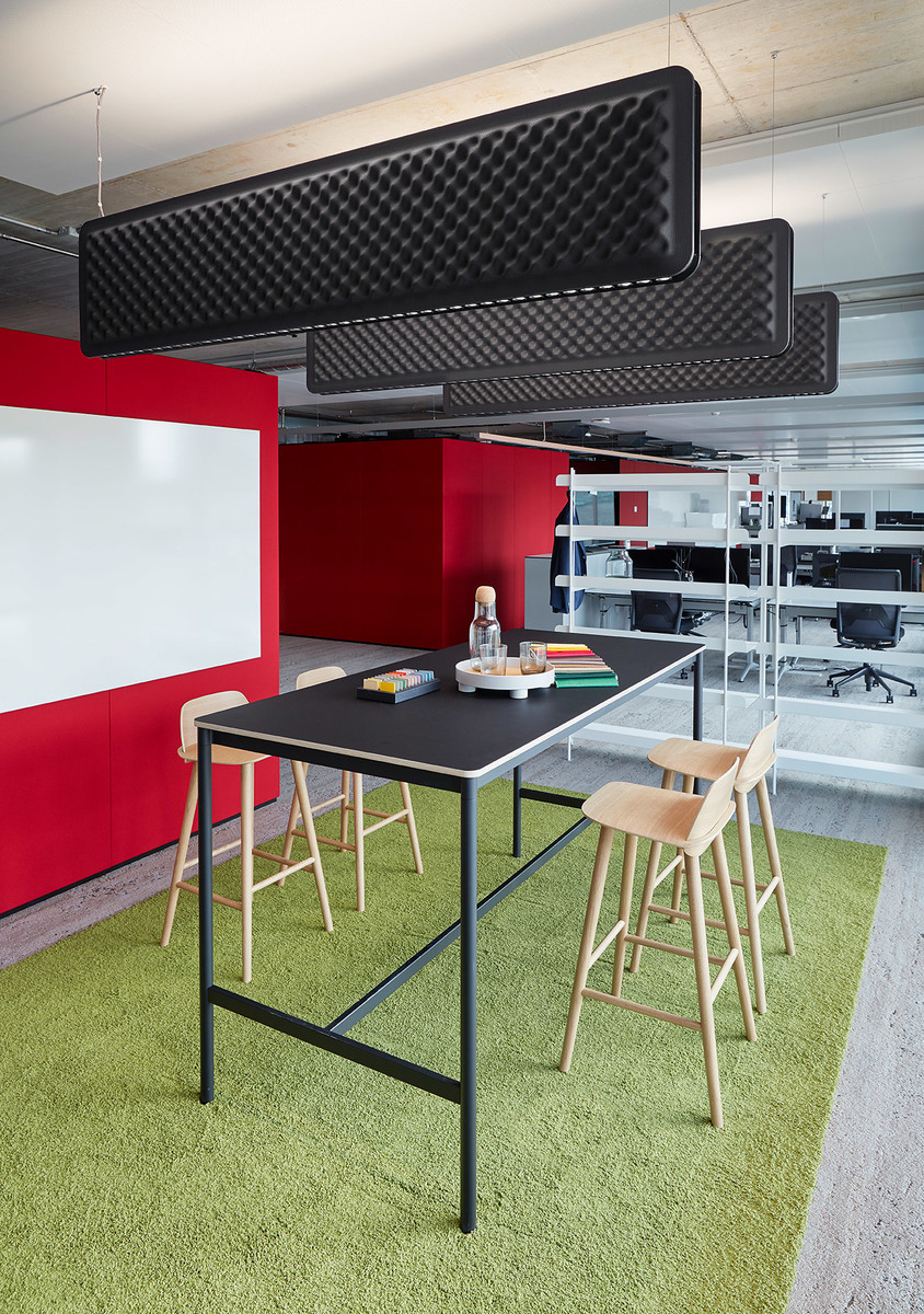 Modern office breakout area with high table, wooden stools, green carpet, red walls, and black acoustic ceiling panels overhead.