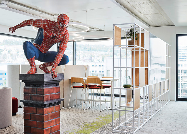 Modern office space with a Spider-Man statue on a brick pedestal, open shelving divider, dining area, and a circular ceiling light fixture.