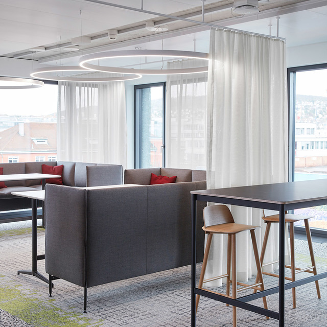 Modern office lounge with Artemide circular ring pendant lights above gray sofas, red cushions, and a high bar table with wooden stools.