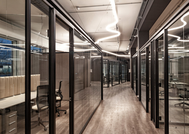 Modern office corridor with glass-partitioned rooms and sinuous white LED pendant lights suspended from an industrial ceiling.